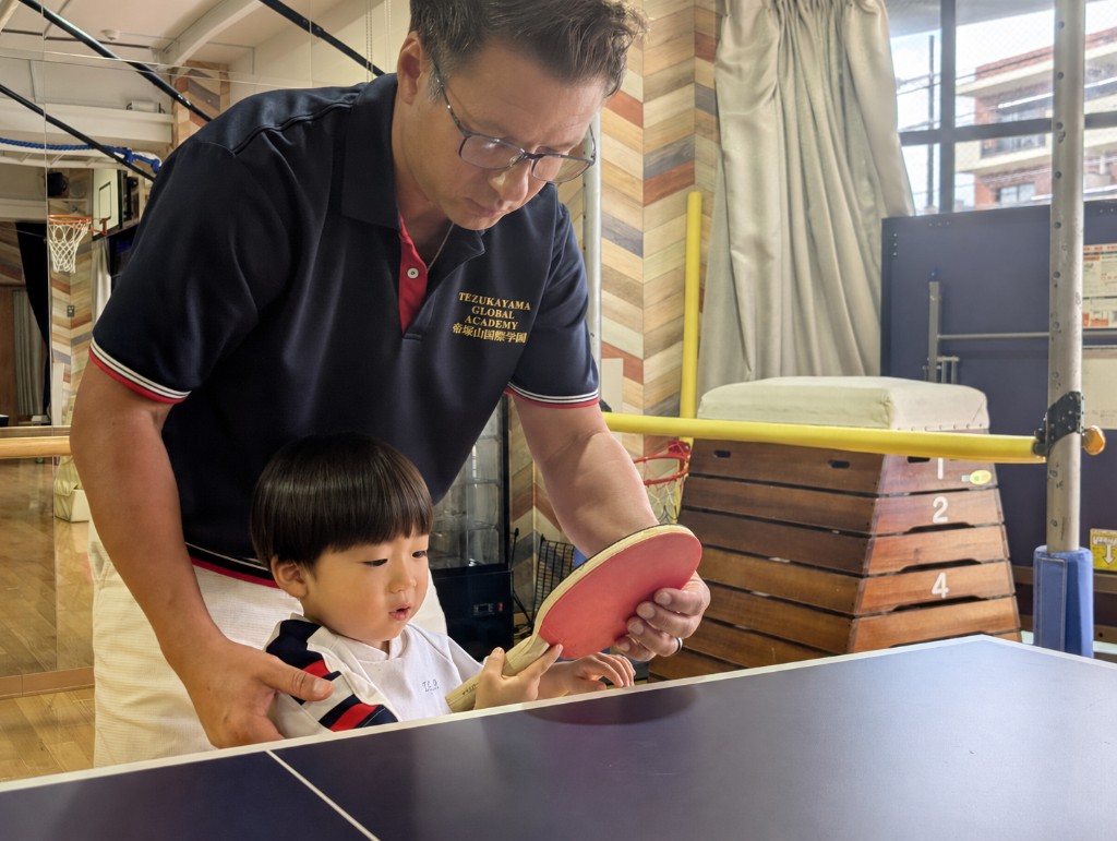 A TGA teacher guiding a young student how to hold a table tennis paddle during PE in the gym