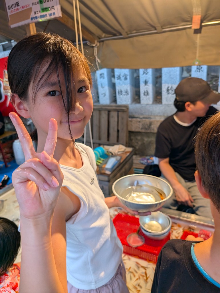 A TGA student holding a goldfish bowl and making a peace sign at a summer festival stall